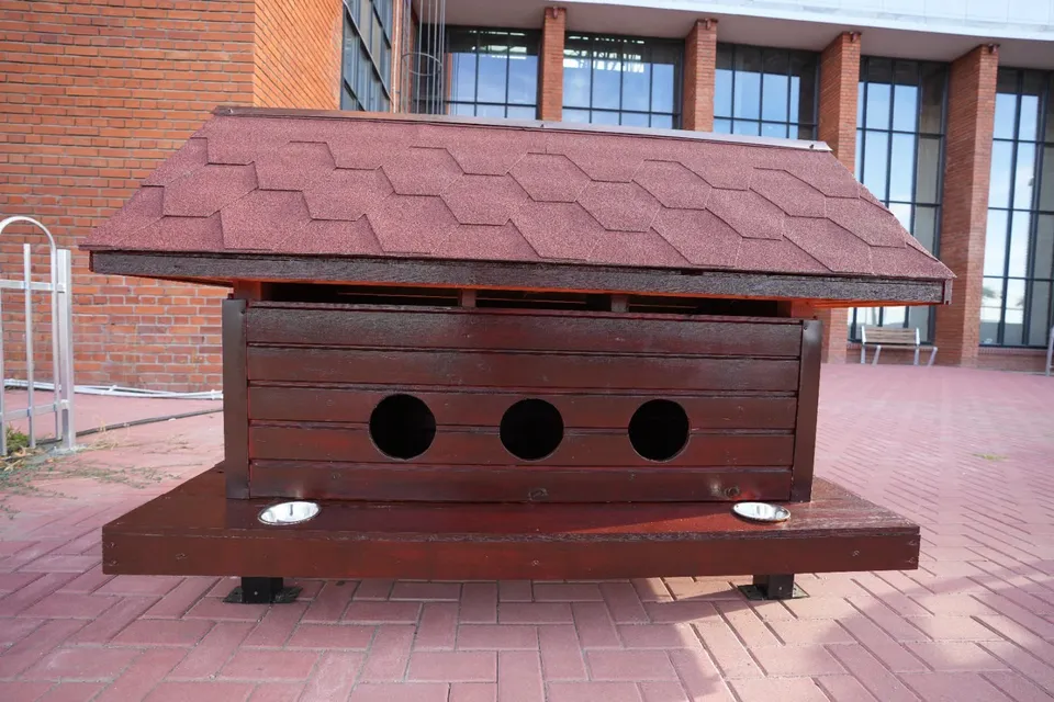 O căsuță din lemn maro cu acoperiș în formă de casă și trei orificii circulare este așezată pe o platformă cu două boluri metalice.