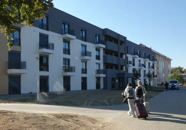 Două persoane cu bagaje merg pe o cărare pavată în fața unui clădire modernă cu ferestre și balcoane negre.
