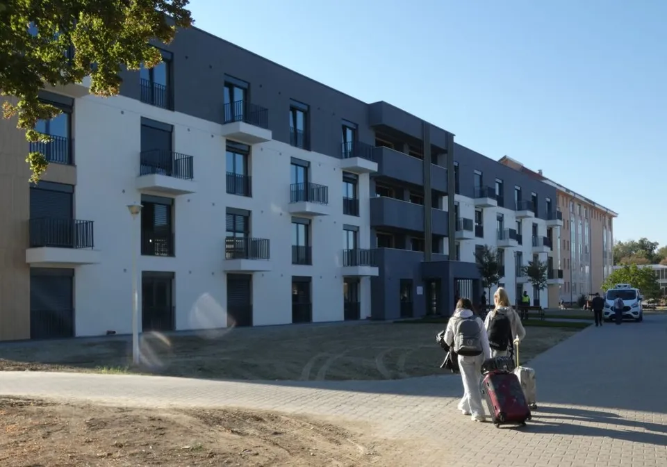 Două persoane cu bagaje merg pe o cărare pavată în fața unui clădire modernă cu ferestre și balcoane negre.