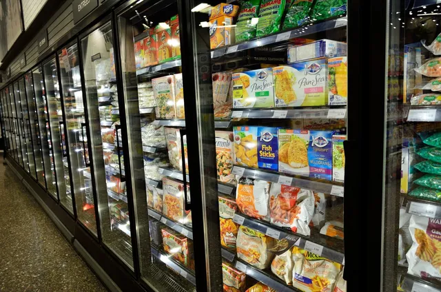 Rânduri de vitrine frigorifice cu uși de sticlă conțin diverse produse alimentare ambalate, expuse pe rafturi în interiorul unui magazin.