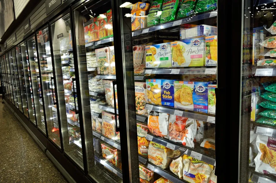Rânduri de vitrine frigorifice cu uși de sticlă conțin diverse produse alimentare ambalate, expuse pe rafturi în interiorul unui magazin.