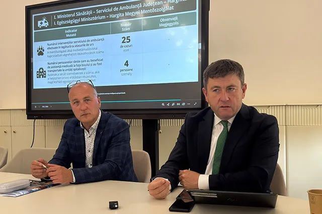 Un bărbat cu sacou și cravată stă la o masă lângă un alt bărbat cu sacou, în fața unui ecran de monitor care afișează un tabel.