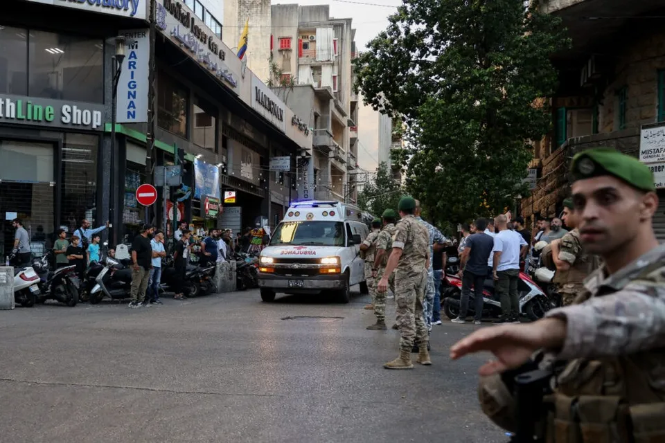 Un grup de bărbați în uniforme militare stă pe un drum urban lângă o ambulanță albă și vehicule parcate.