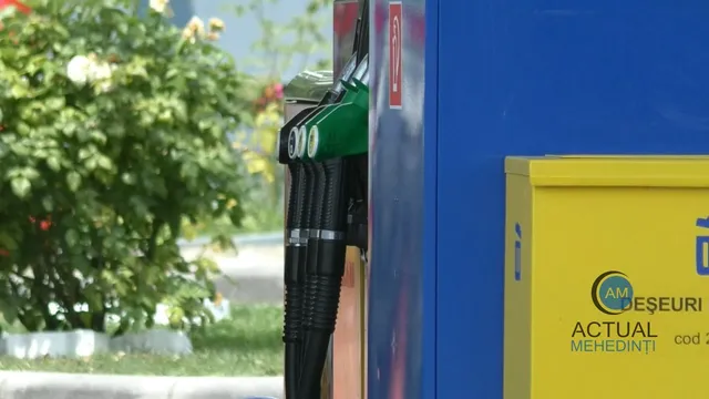 Două duze de pompat combustibil negre cu accesorii verzi sunt suspendate pe o stație de alimentare lângă un recipient galben.