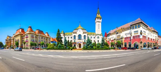 Clădiri colorate cu acoperișuri în formă de turn se află pe o stradă lângă un spațiu verde sub un cer albastru.
