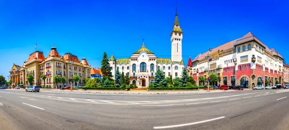 Clădiri colorate cu acoperișuri în formă de turn se află pe o stradă lângă un spațiu verde sub un cer albastru.