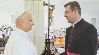 Un bărbat cu pălărie albă și un bărbat cu haine negre cu detalii roz stau față în față lângă un crucifix înalt într-o încăpere luminoasă.