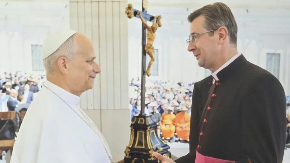 Un bărbat cu pălărie albă și un bărbat cu haine negre cu detalii roz stau față în față lângă un crucifix înalt într-o încăpere luminoasă.