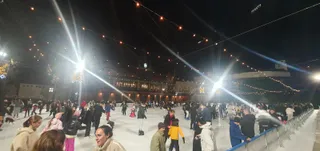 O patinoare artificială aglomerată, luminată de instalații puternice de iluminat și șiruri de lumini decorative suspendate în aer, cu multe persoane patinând pe gheață.