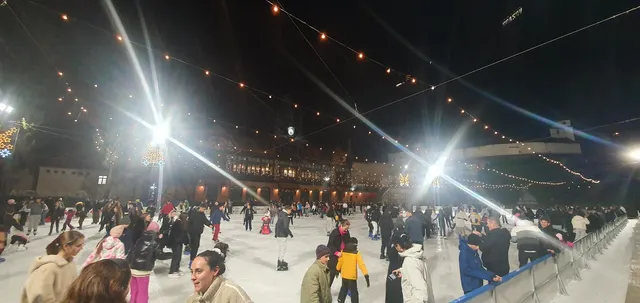 O patinoare artificială aglomerată, luminată de instalații puternice de iluminat și șiruri de lumini decorative suspendate în aer, cu multe persoane patinând pe gheață.