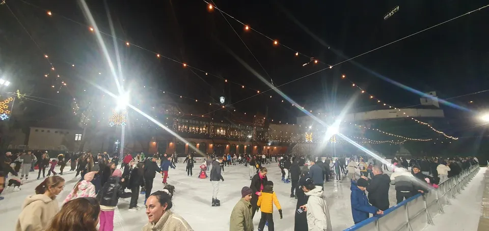 O patinoare artificială aglomerată, luminată de instalații puternice de iluminat și șiruri de lumini decorative suspendate în aer, cu multe persoane patinând pe gheață.