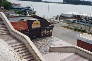 O vedere de sus asupra unor trepte de piatră, unui stand de vânzare negru cu desene și unei porți de lemn spre un fluviu.