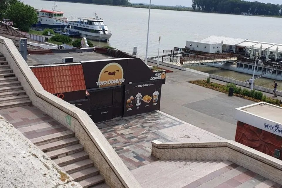 O vedere de sus asupra unor trepte de piatră, unui stand de vânzare negru cu desene și unei porți de lemn spre un fluviu.