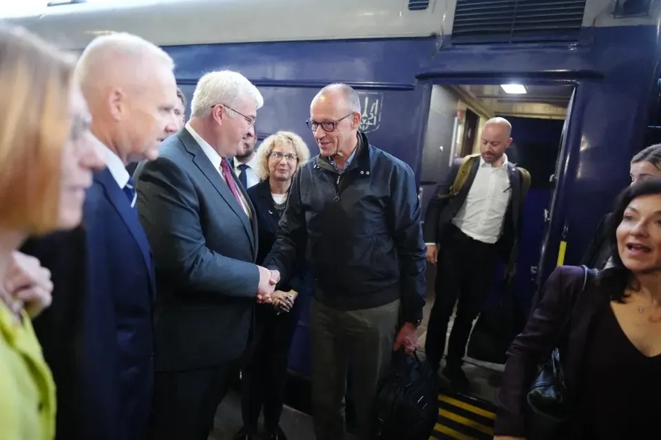 Un grup de oameni în haine formale stau lângă o intrare de tren albastră și neagră, în timp ce un bărbat cu ochelari coboară din vagon.