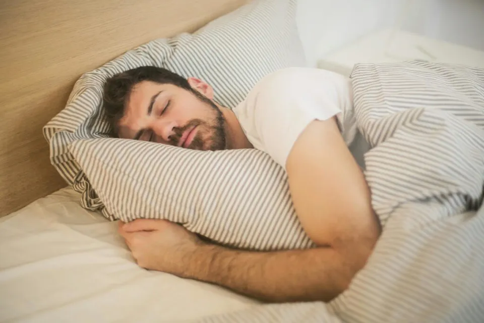 Un bărbat cu barbă doarme pe o pernă cu dungi sub o pătură în timp ce se află în pat.