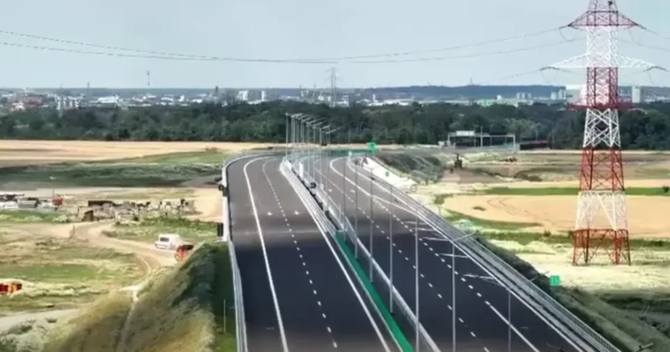 O porțiune de autostradă cu mai multe benzi de rulare, marcaje albe și o linie verde centrală, trece pe lângă un turn de înaltă tensiune.