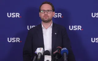Un bărbat cu ochelari și sacou în fața unui fundal albastru pe care apare textul USR, stând în spatele mai multor microfoane.