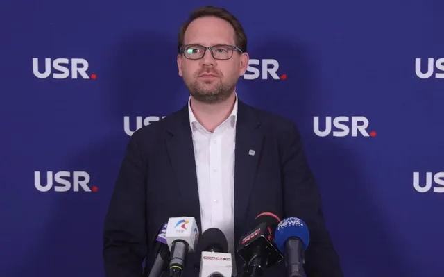 Un bărbat cu ochelari și sacou în fața unui fundal albastru pe care apare textul USR, stând în spatele mai multor microfoane.