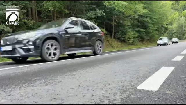 Un autoturism gri circulă pe un drum asfaltat cu linii albe marcate, având copaci și alte vehicule în fundal.