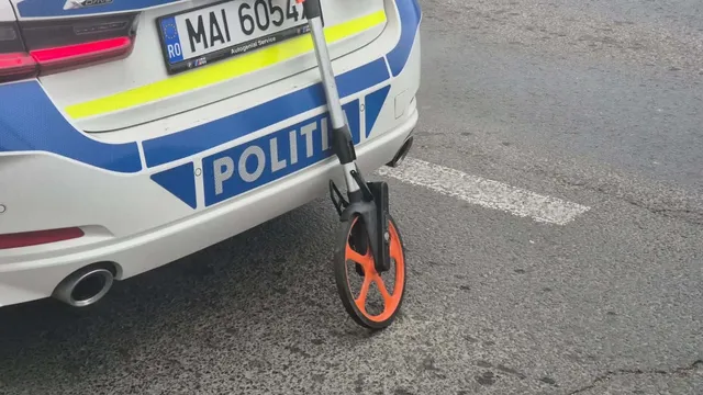 Un trotinet cu roți portocalii este oprit lângă partea din spate a unei mașini de poliție albe cu marcaje albastre și galbene.