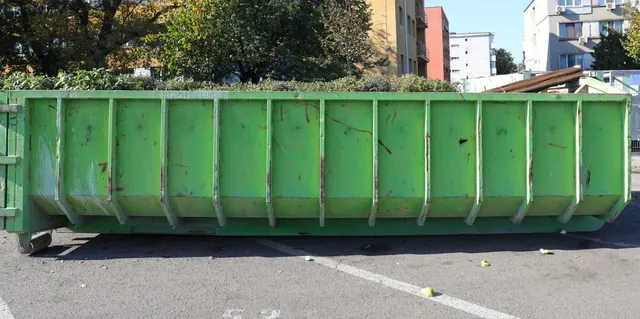 Un container metalic de culoare verde este poziționat pe un asfalt gri lângă un gard de vegetație și clădiri.