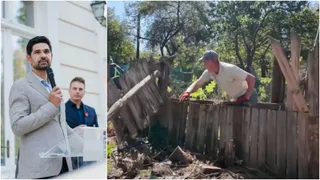 Un bărbat cu cămașă deschisă și pantaloni scurți se apleacă peste un gard de lemn improvizat într-o zonă cu vegetație.