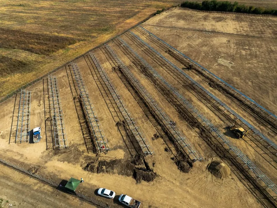 O vedere aeriană prezintă mai multe rânduri de structuri metalice lungi instalate pe un teren de construcții lângă două vehicule albe.