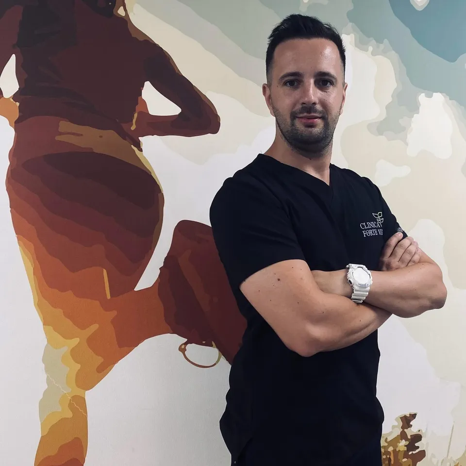 Un bărbat cu părul scurt și barbă poartă un tricou negru și un ceas alb, stând cu brațele încrucișate în fața unui perete decorat.