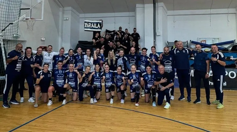 O echipă de femei în echipamente sportive albastre și negre pozează împreună pe un teren de sport în interiorul unei săli.