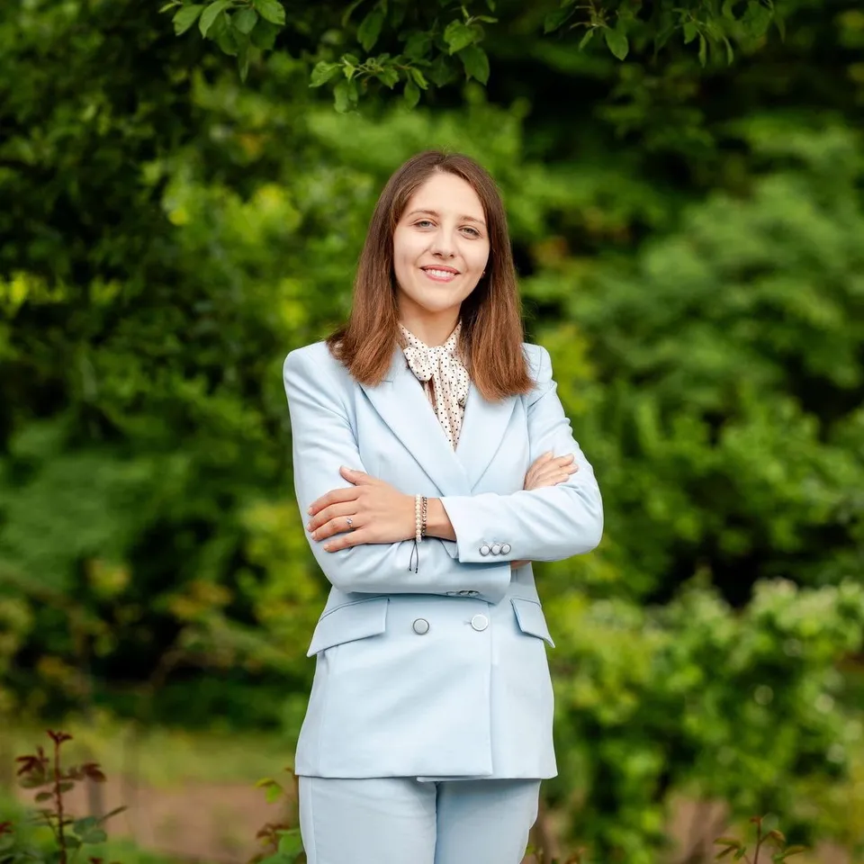 O femeie cu părul lung și deschis la culoare poartă un costum albastru deschis și stă cu brațele încrucișate în fața unor plante verzi.