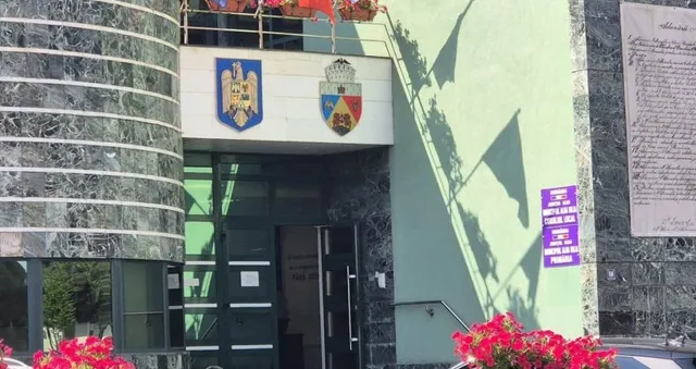O clădire cu fațadă de marmură gri și verde prezintă două embleme colorate deasupra unei intrări cu uși de sticlă și cadre negre.