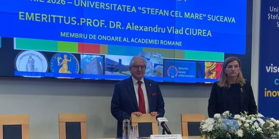 Un bărbat cu ochelari și cravată roșie stă lângă o femeie în fața unui ecran albastru pe care scrie text galben și alb.