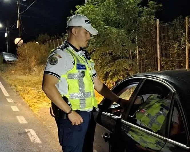 Un agent de poliție în uniformă și vestă reflectorizantă stă lângă o mașină neagră pe un drum asfaltat noaptea.