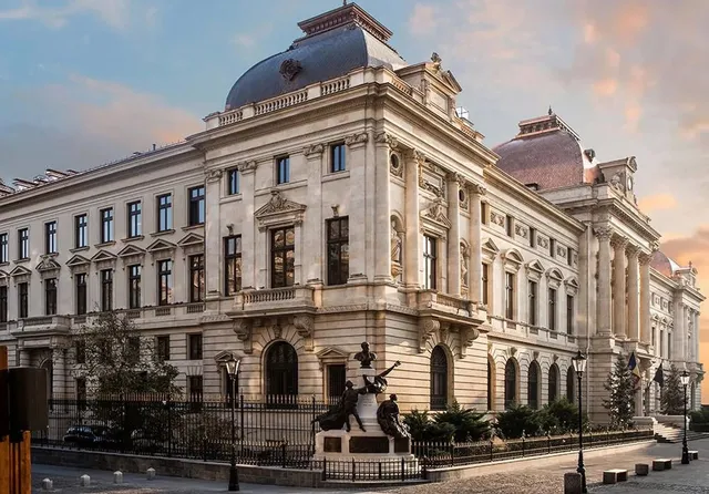 O clădire monumentală de piatră cu ferestre înalte, coloane și un cupolă gri, situată lângă un trotuar cu un monument sculptat.