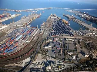 O vedere aeriană a unui port cu nave, macarale, containere colorate și infrastructură feroviară lângă o apă de culoare albastră.