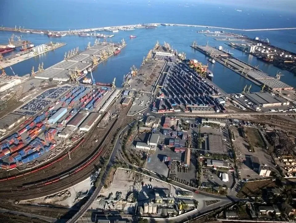 O vedere aeriană a unui port cu nave, macarale, containere colorate și infrastructură feroviară lângă o apă de culoare albastră.