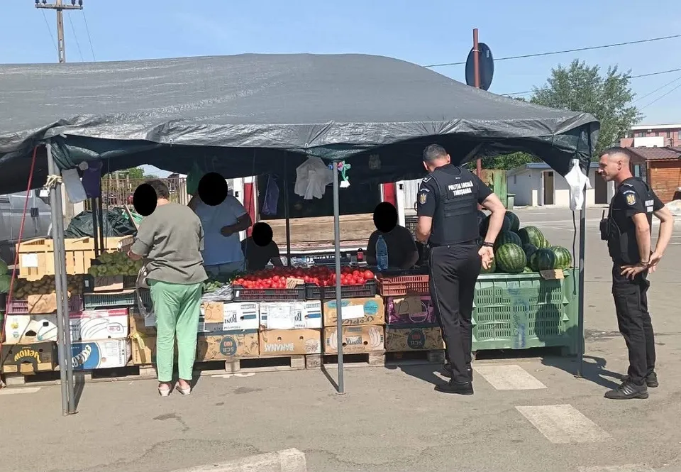 Unii bărbați stau lângă un tarab de fructe și legume acoperit de un copertină neagră, în prezența unor persoane în uniformă întunecată.