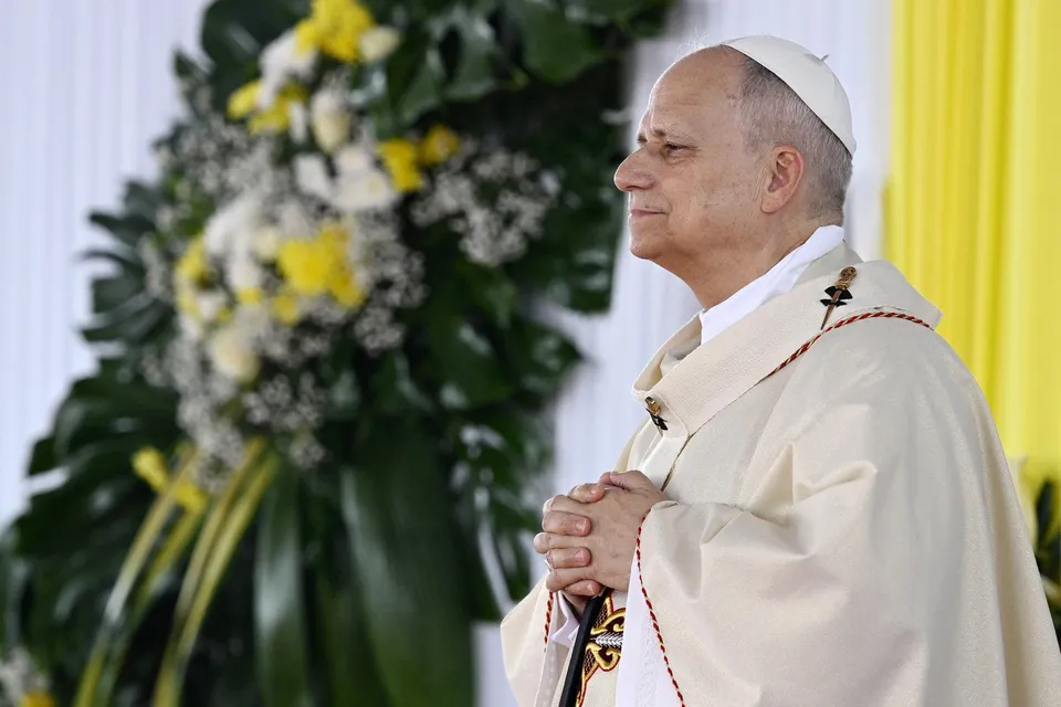 Un bărbat în vârstă cu zucchetto alb și robă albă stă cu mâinile împreunate în fața unui buchet de flori galbene și verzi.