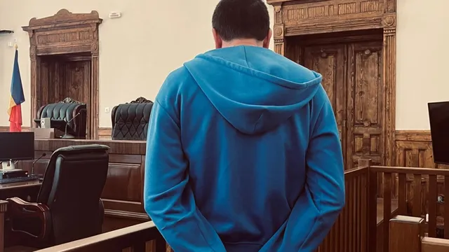 Un bărbat cu un hanorac albastru stă cu spatele la cameră într-o încăpere cu mobilier din lemn și scaune de birou.