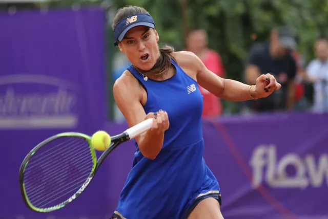 O femeie cu visieră albastră și echipament sportiv albastru lovește o minge de tenis galbenă cu o rachetă.