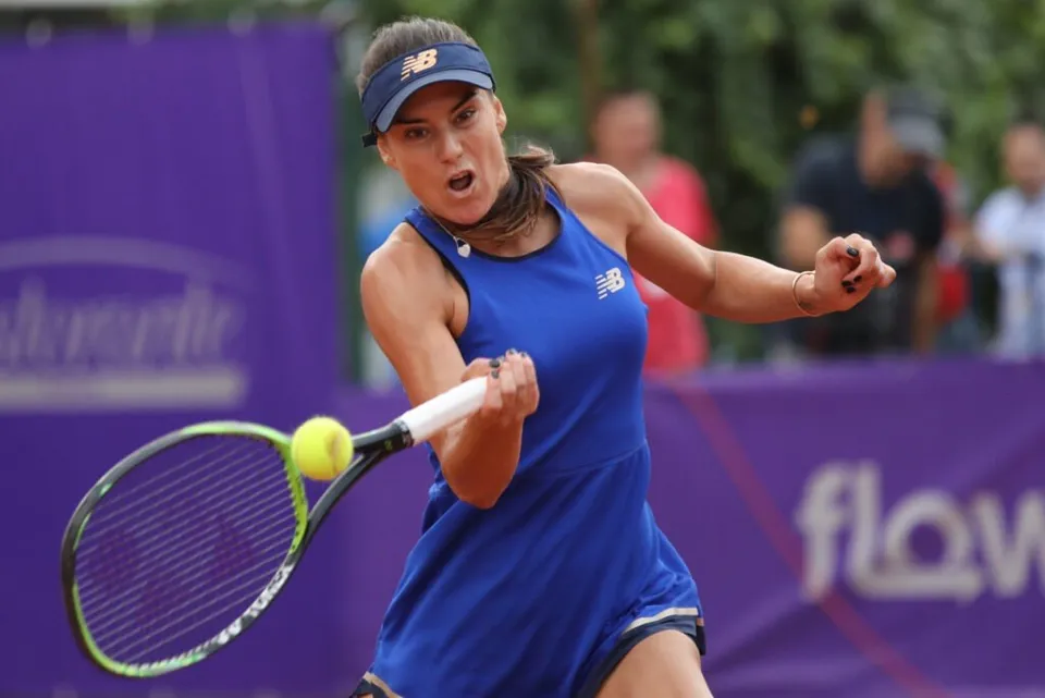 O femeie cu visieră albastră și echipament sportiv albastru lovește o minge de tenis galbenă cu o rachetă.