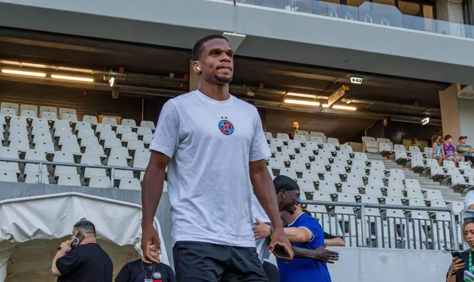 Un bărbat cu tricou alb și căști albe stă în picioare în fața unor scaune albe de stadion, în timp ce alți oameni sunt vizibili în fundal.