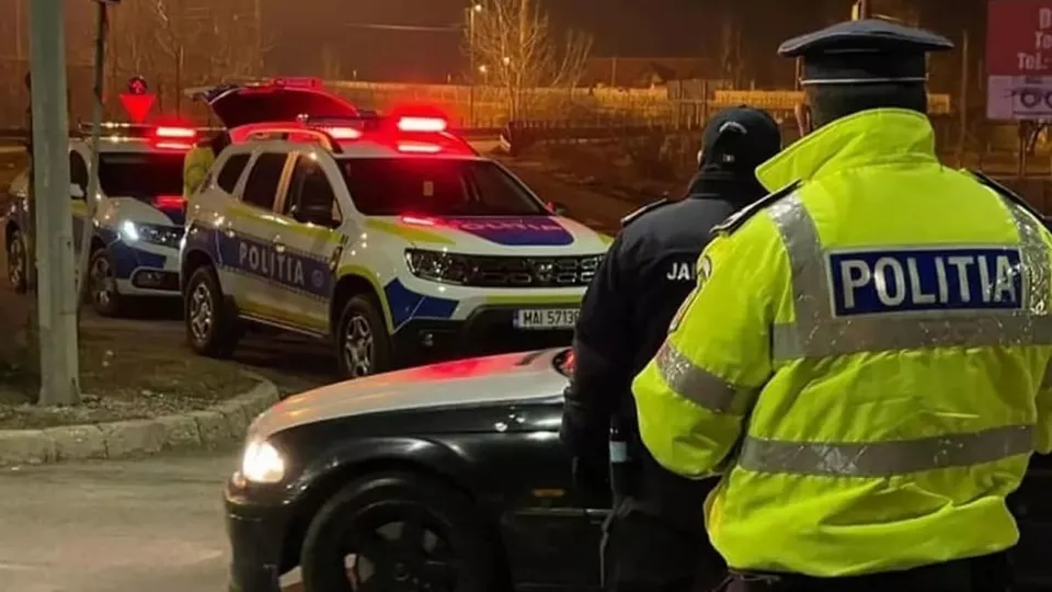 Un polițist în uniformă galbenă reflectorizantă stă lângă un vehicul negru, în timp ce două mașini de poliție cu girofare aprinse sunt parcate în fundal.