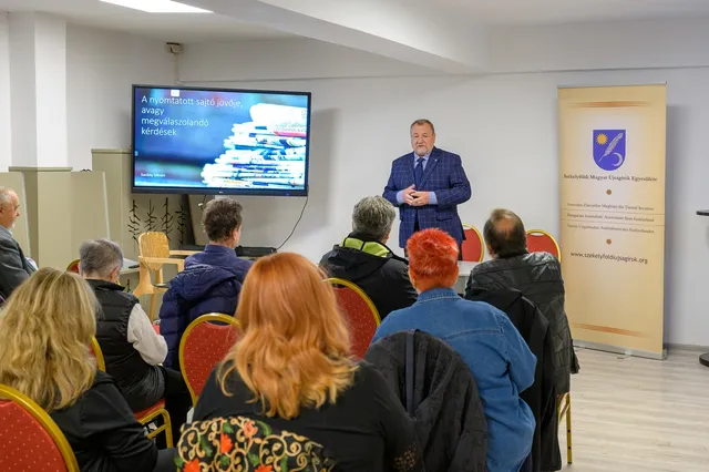 Un bărbat într-un costum albastru stă în fața unui grup de persoane așezate pe scaune într-o încăpere cu un ecran și un panou vertical.