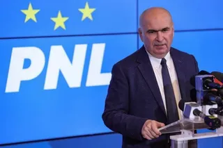 Un bărbat cu sacou închis și cravată stă în fața unui panou albastru cu literele PNL și vorbeste la microfoane.