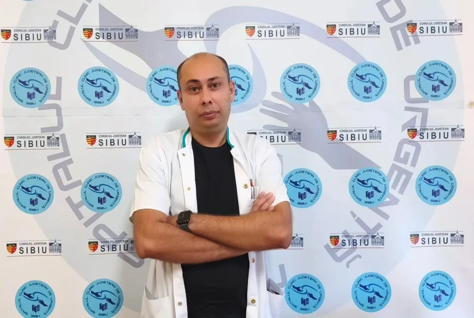 Un bărbat cu halat alb și brațele încrucișate stă în fața unui fundal alb cu diverse embleme circulare și dreptunghiulare albastre și colorate.