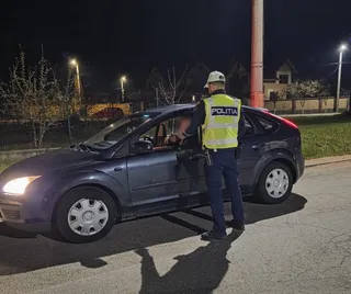 Un agent de poliție cu vestă reflectorizantă stă lângă portiera deschisă a unei mașini gri pe un drum asfaltat noaptea.