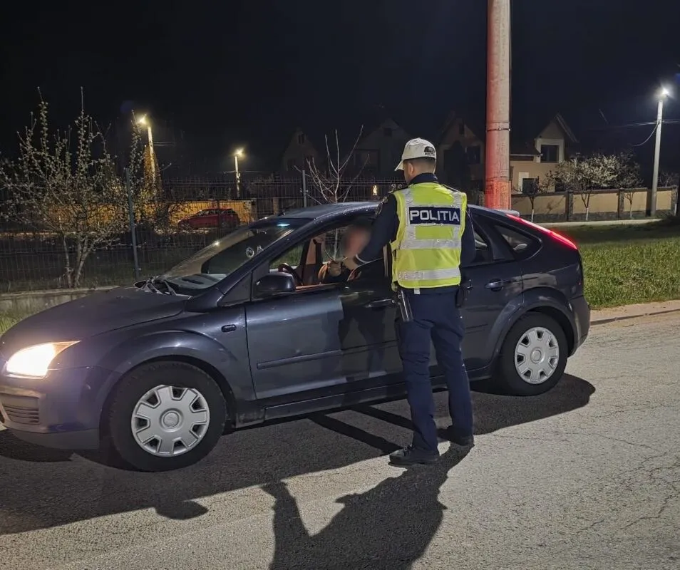 Un agent de poliție cu vestă reflectorizantă stă lângă portiera deschisă a unei mașini gri pe un drum asfaltat noaptea.