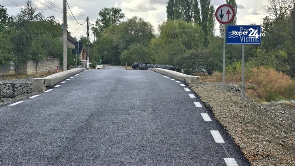 O porțiune de drum asfaltat cu linii albe marcate pe margini trece peste o structură de pod înconjurat de vegetație și un semn de circulație.