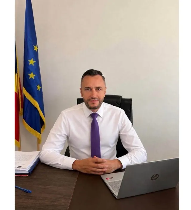 Un bărbat cu cămașă albă și cravată mov stă la un birou lângă un laptop și două steaguri.